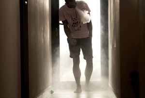 A Young Man Of African-American Descent Is Standing In A Hallway, Blowing Vapour Of His E-Cigarette