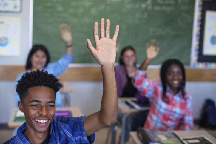 Smiling diverse multi-cultural students raising hands in classroom