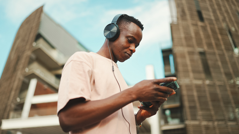 Young african student sitting outside of university wearing headphones, using phone, listening to music