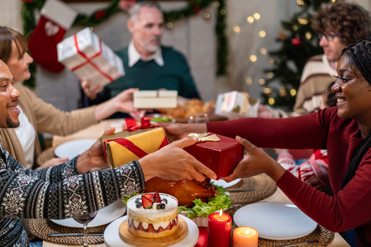 Multi-ethnic family exchanging presents during Christmas party at home. Attractive diverse people receive gift from relatives while having dinner celebrate holiday Thanksgiving on dining table.